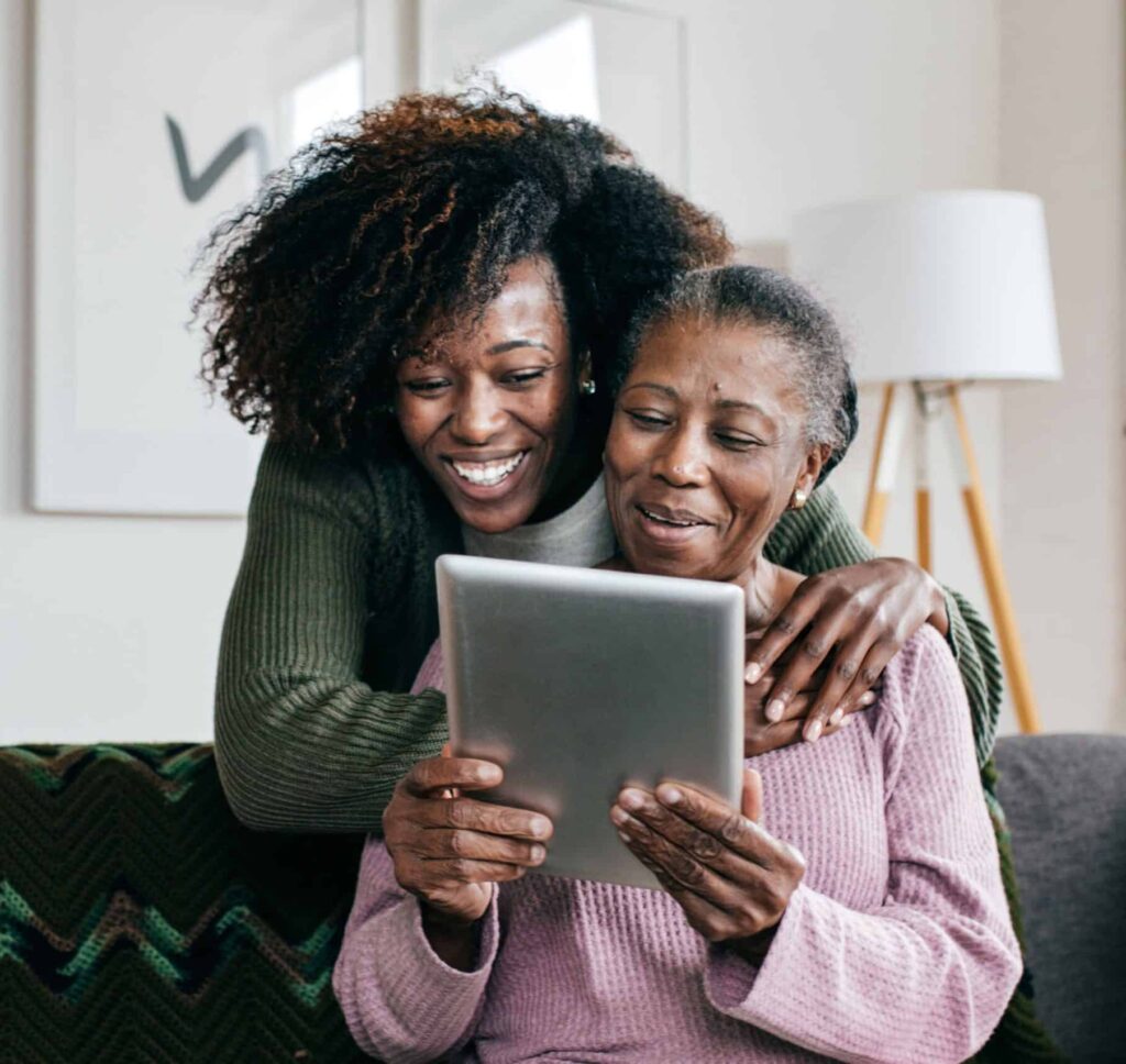 Two women sitting on a couch, sharing a tablet and smiling at something they see together.